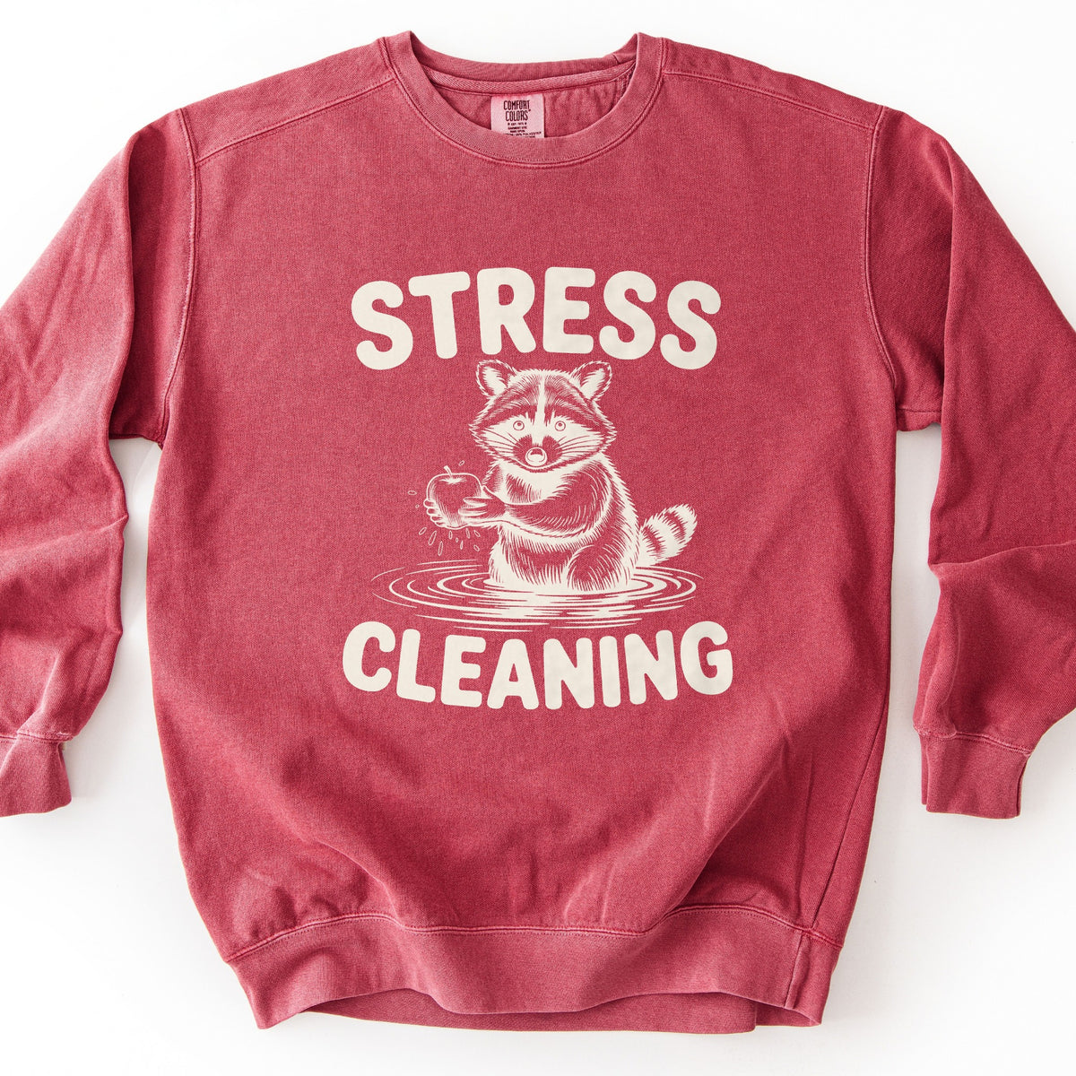 Stress Cleaning Comfort Colors crewneck sweatshirt in Crimson, laid flat. Cream print of a sketched raccoon standing in rippling water, washing an apple, between the words STRESS and CLEANING. The sweatshirt lies on a white surface.