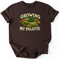 Growing My Palette Comfort Colors T-shirt in Dark Chocolate, laid flat. An artist's wooden palette and paintbrush covered in green moss and plants, between the words Growing My Palette in cream letters edged with moss. The sleeves are rolled on a white background.