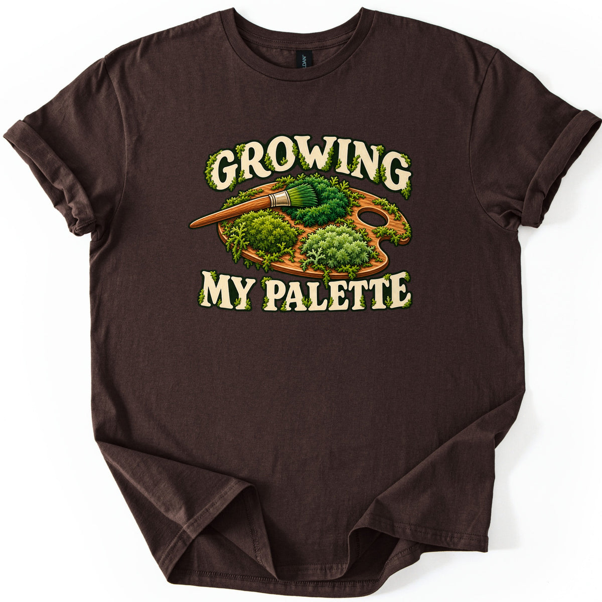 Growing My Palette Comfort Colors T-shirt in Dark Chocolate, laid flat. An artist's wooden palette and paintbrush covered in green moss and plants, between the words Growing My Palette in cream letters edged with moss. The sleeves are rolled on a white background.