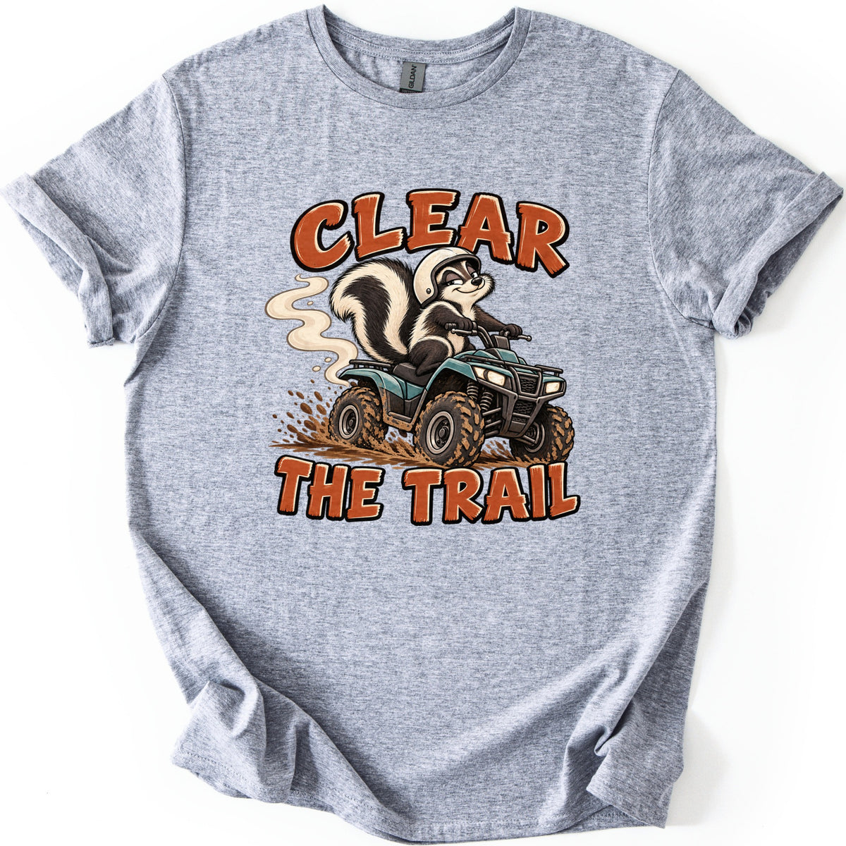 Clear the Trail Comfort Colors T-shirt in Sport Grey, laid flat. A cartoon skunk in a white helmet rides a teal ATV, kicking up mud and a swirl of smoke, between the words Clear The Trail in rust-orange letters. The shirt lies on a white background with cuffed sleeves.