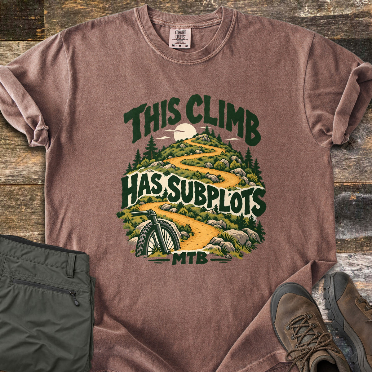 This Climb Has Subplots Comfort Colors T-shirt in Espresso, laid flat. A winding dirt trail climbs a rocky, pine-covered hill under a pale sun, with a mountain bike wheel in front, and the words This Climb Has Subplots MTB. Olive pants and brown hiking boots sit on weathered wood.