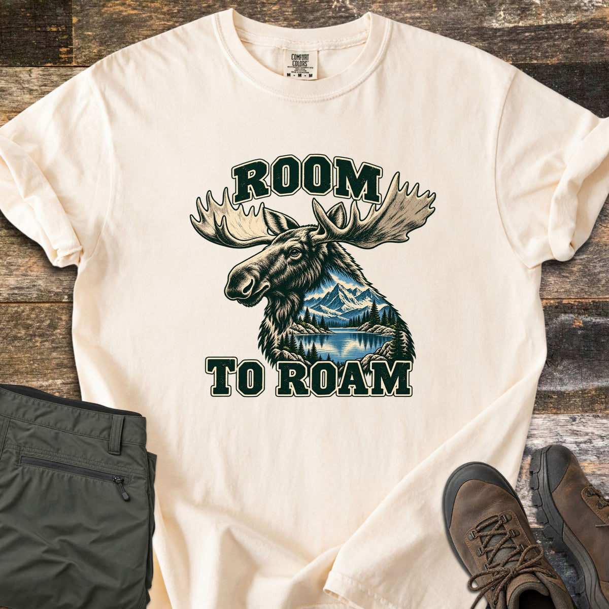 Room to Roam Comfort Colors T-shirt in Ivory, laid flat. A moose head with large antlers, a mountain lake scene inside its neck, between the words Room To Roam in dark green. Olive hiking pants and brown hiking boots sit beside it on weathered wood planks.
