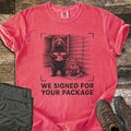 We Signed For Your Package Comfort Colors T-shirt in Watermelon, laid flat. A black-ink sketch shows a bear holding an open box beside a seated fox on a log cabin porch, framed by camera corner marks above the words We Signed For Your Package. Olive pants and brown hiking boots sit on wood planks.
