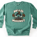 Mud Magnet Comfort Colors crewneck sweatshirt in Light Green, laid flat. Cream block letters read Mud Magnet around a dark horseshoe magnet pulling brown mud splashes toward it, above a rider on an ATV churning through mud. Shown on a white background.