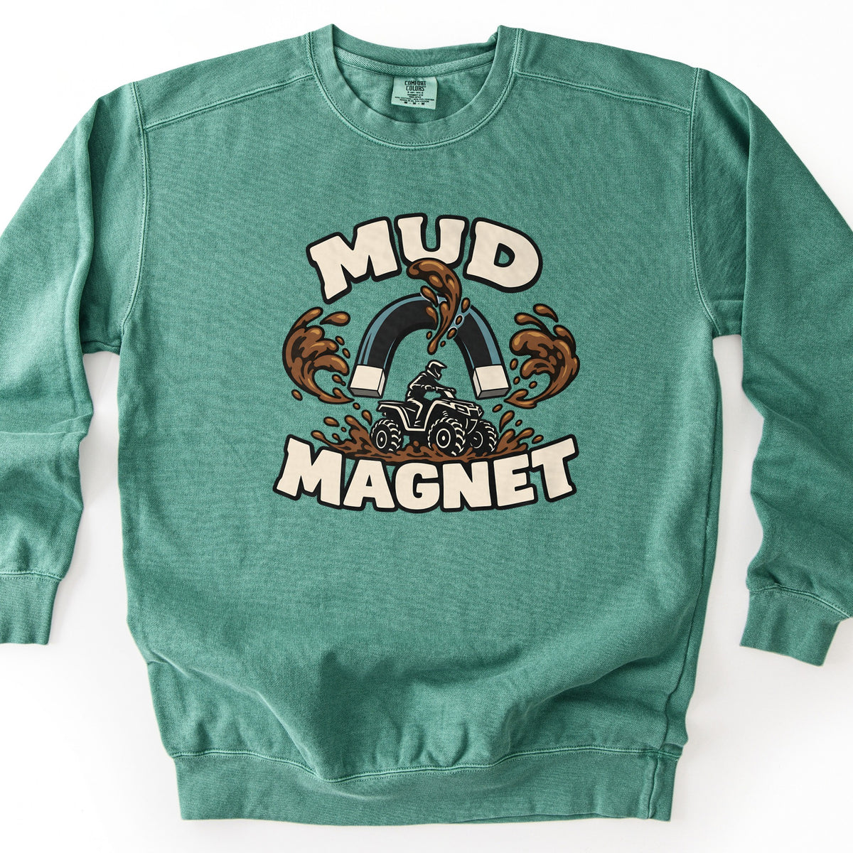 Mud Magnet Comfort Colors crewneck sweatshirt in Light Green, laid flat. Cream block letters read Mud Magnet around a dark horseshoe magnet pulling brown mud splashes toward it, above a rider on an ATV churning through mud. Shown on a white background.