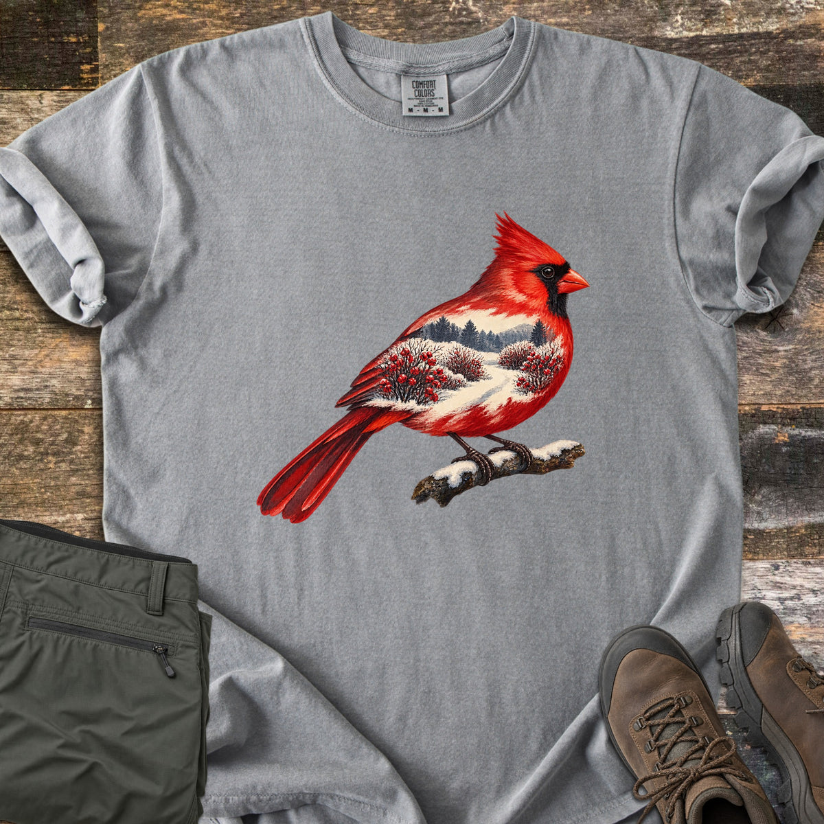 Winter Cardinal Comfort Colors T-shirt in Grey, laid flat. A red cardinal perched on a snowy branch, its body filled with a snowy landscape of pine trees and red berry bushes. Rolled sleeves on weathered wood, with olive pants and brown hiking boots beside it.