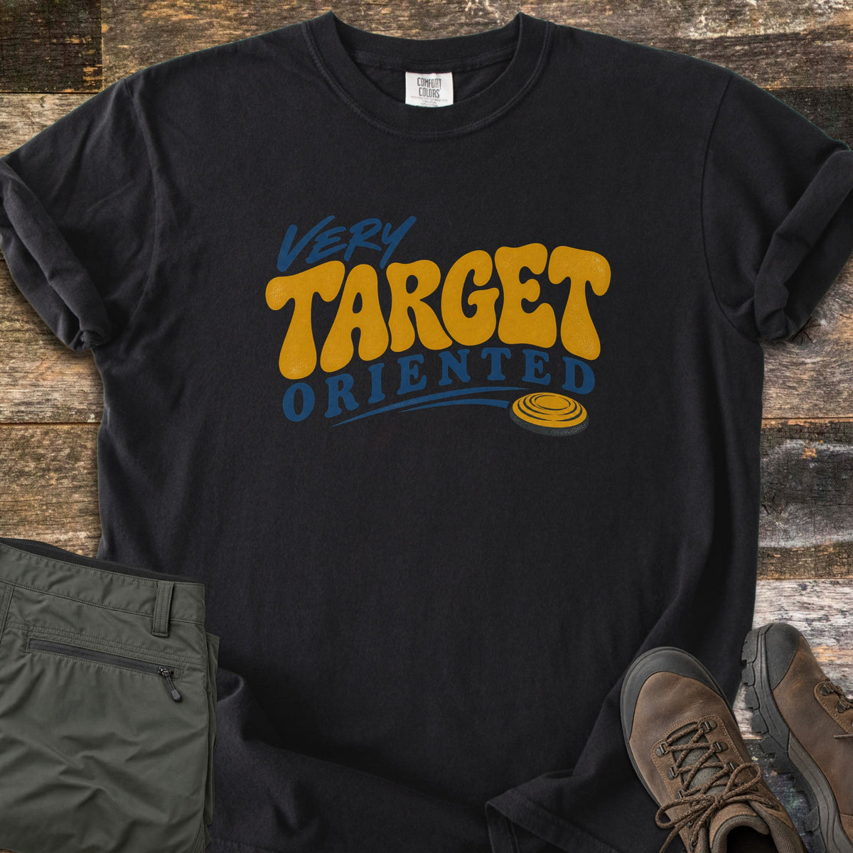 Very Target Oriented Comfort Colors T-shirt in Black, laid flat. The words Very Target Oriented in blue and gold retro lettering, with a gold clay pigeon target trailing a blue swoosh. It lies on weathered wood beside olive cargo pants and brown hiking boots.