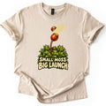 Small Moss Big Launch Comfort Colors T-shirt in Sand, laid flat. An orange moss capsule on a curved stalk rises from leafy green moss, its lid popping off as yellow spores burst out, above the words Small Moss Big Launch. Rolled sleeves, on a white background.