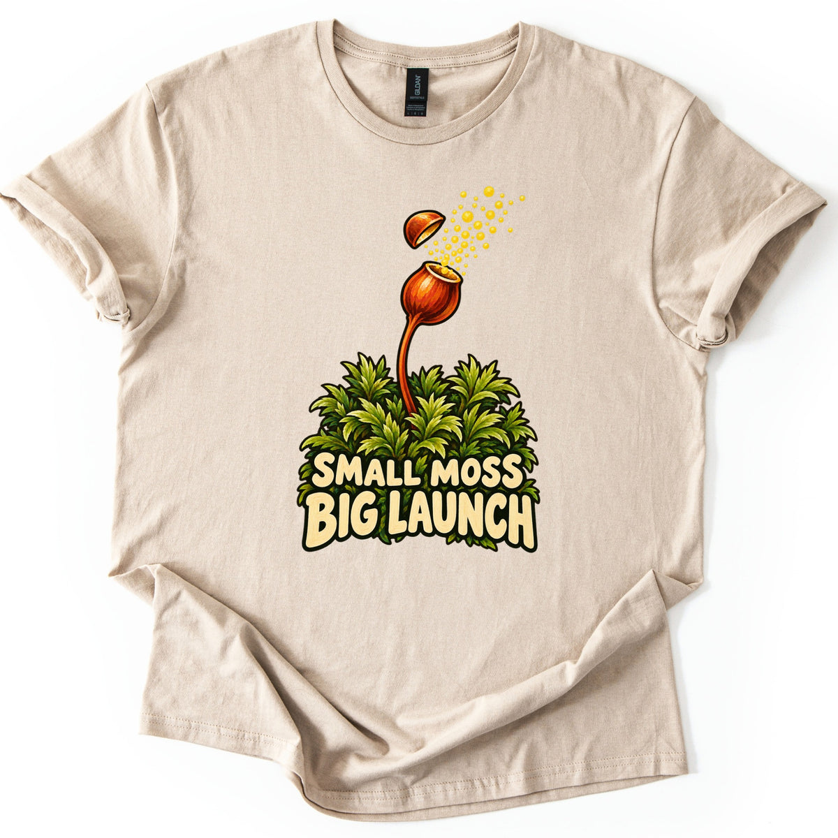 Small Moss Big Launch Comfort Colors T-shirt in Sand, laid flat. An orange moss capsule on a curved stalk rises from leafy green moss, its lid popping off as yellow spores burst out, above the words Small Moss Big Launch. Rolled sleeves, on a white background.