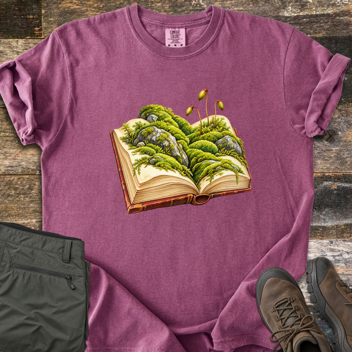 Moss Storybook Comfort Colors T-shirt in Berry, laid flat. An illustrated open book with green moss, gray rocks, and tall moss sporophytes growing from its pages. Rolled sleeves, on weathered wood with folded olive pants and brown hiking boots.
