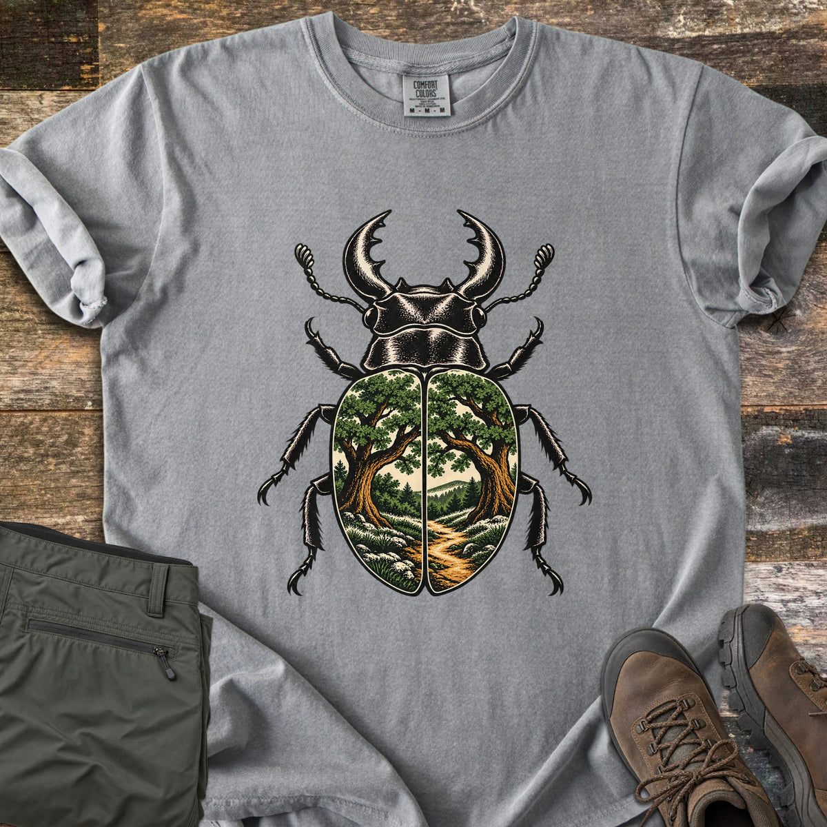Beetle Forest Path Comfort Colors T-shirt in Grey, laid flat. A black stag beetle whose wing covers show a forest scene of two oak trees framing a dirt path toward distant hills. Folded olive hiking pants and brown hiking boots sit beside it on weathered wood planks.