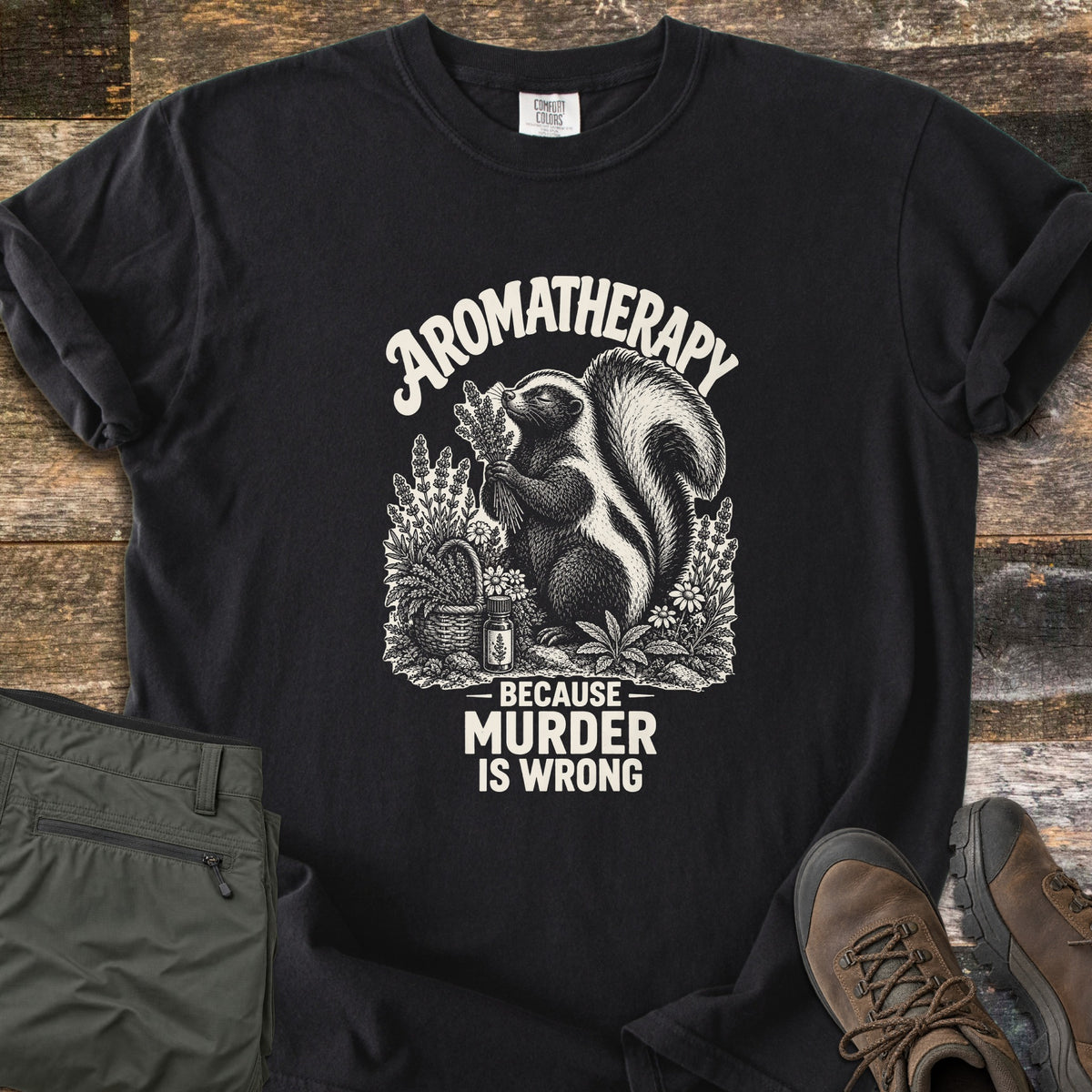 Aromatherapy Because Murder Is Wrong Comfort Colors T-shirt in Black, laid flat. An engraving-style skunk sniffs a bundle of lavender among flowers, a basket and an oil bottle, in cream ink, with the words Aromatherapy, Because Murder Is Wrong. Olive pants and brown hiking boots lie on wood planks.