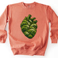 Moss Heart Comfort Colors crewneck sweatshirt in Terracotta, laid flat. The front shows an anatomical heart made of green moss clumps with pale cream tufts and tiny sprouts, its three vessels open at the top. No words are printed. It lies on a white surface, sleeves at its sides.