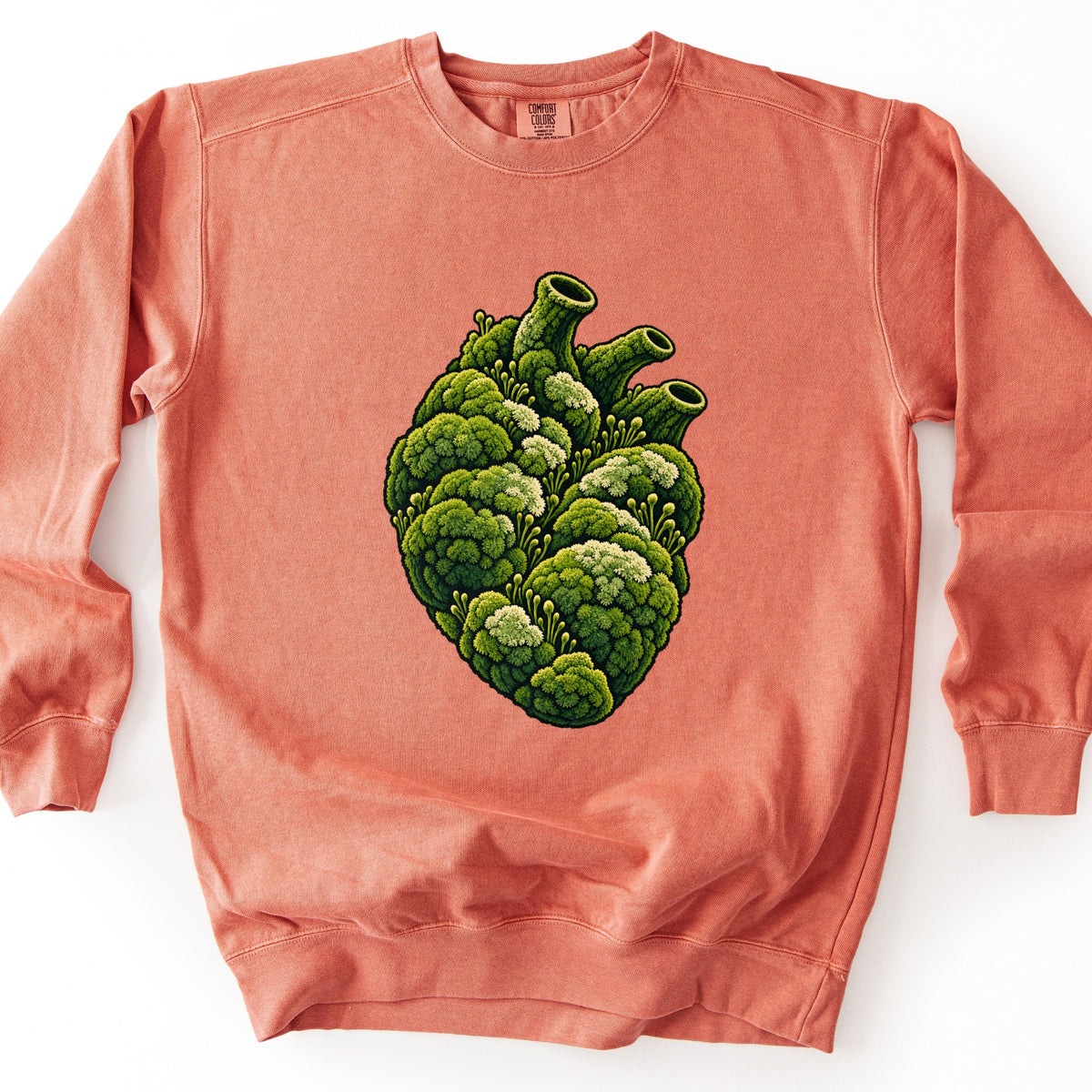 Moss Heart Comfort Colors crewneck sweatshirt in Terracotta, laid flat. The front shows an anatomical heart made of green moss clumps with pale cream tufts and tiny sprouts, its three vessels open at the top. No words are printed. It lies on a white surface, sleeves at its sides.