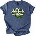 Ask Me About Moss Comfort Colors T-shirt in Heather Navy, laid flat. A cream speech bubble reads ASK ME ABOUT MOSS in bold dark green letters above a cartoon clump of green moss with three stalked spore capsules. The shirt has rolled sleeves on a white background.