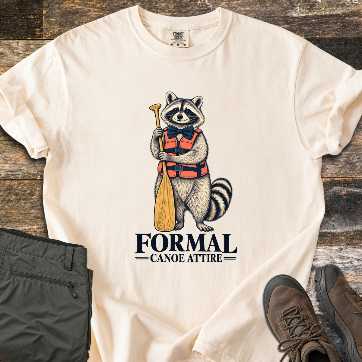 Formal Canoe Attire Comfort Colors T-shirt in Ivory, laid flat. A raccoon in an orange life vest and navy bow tie stands holding a wooden canoe paddle above the words Formal Canoe Attire. Olive hiking pants and brown hiking boots sit beside it on weathered wood planks.