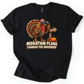 Migration Plans Changed for November Comfort Colors T-shirt in Black, laid flat. A cartoon turkey looks through binoculars beside a wooden arrow sign and a small pumpkin, above the words Migration Plans Changed for November. The shirt has rolled sleeves on a white background.