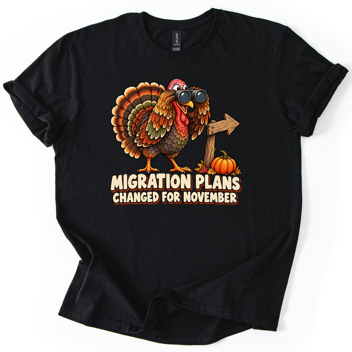Migration Plans Changed for November Comfort Colors T-shirt in Black, laid flat. A cartoon turkey looks through binoculars beside a wooden arrow sign and a small pumpkin, above the words Migration Plans Changed for November. The shirt has rolled sleeves on a white background.