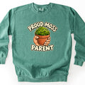 Proud Moss Parent Comfort Colors crewneck sweatshirt in Light Green, laid flat. A hand holds a terracotta pot of smiling cartoon moss between the words Proud Moss and Parent in cream bubble letters. The sweatshirt lies on a plain white background.