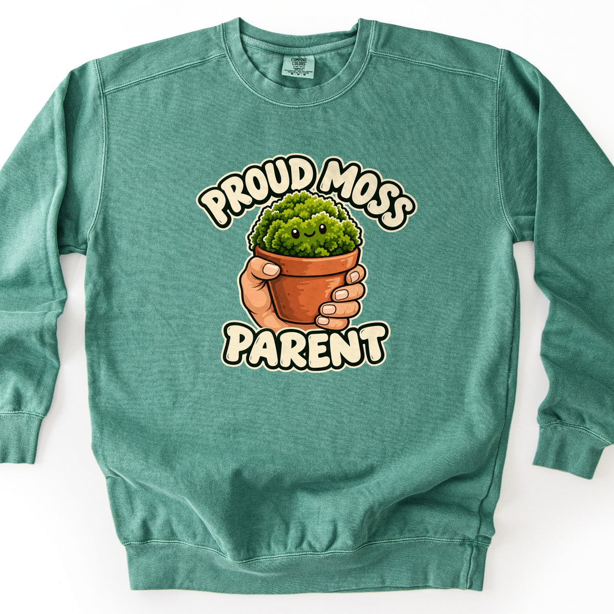 Proud Moss Parent Comfort Colors crewneck sweatshirt in Light Green, laid flat. A hand holds a terracotta pot of smiling cartoon moss between the words Proud Moss and Parent in cream bubble letters. The sweatshirt lies on a plain white background.