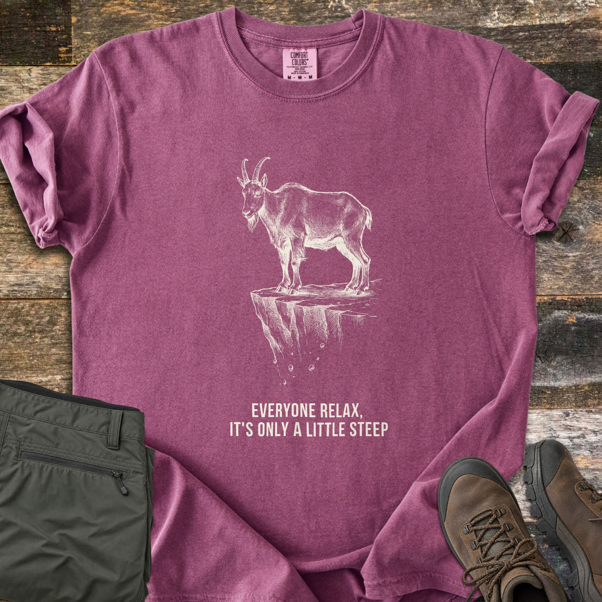 Everyone Relax It's Only A Little Steep Comfort Colors T-shirt in Berry, laid flat. A white sketch of a horned mountain goat standing on a crumbling cliff ledge above the words Everyone Relax, It's Only A Little Steep. Sleeves are rolled, on wood planks with green hiking pants and brown boots.