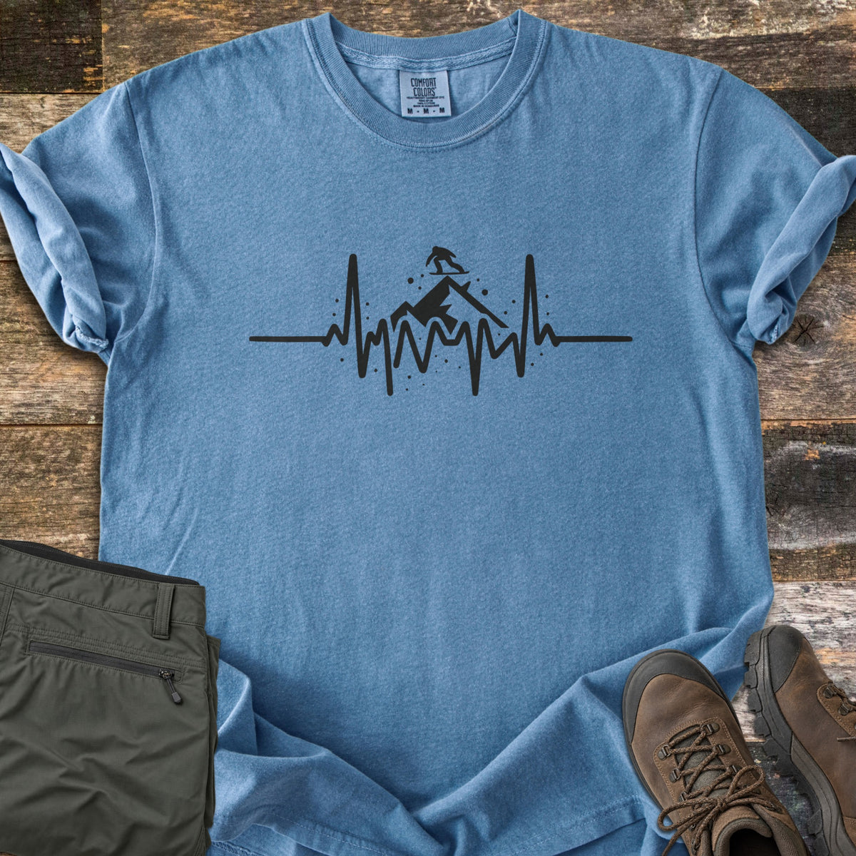Snow Boarder Heartbeat Comfort Colors T-shirt in Blue Jean, laid flat. A black heartbeat line rises into a mountain peak with a snowboarder riding on top, surrounded by snow dots. Folded olive hiking pants and brown leather boots sit beside it on weathered wood planks.
