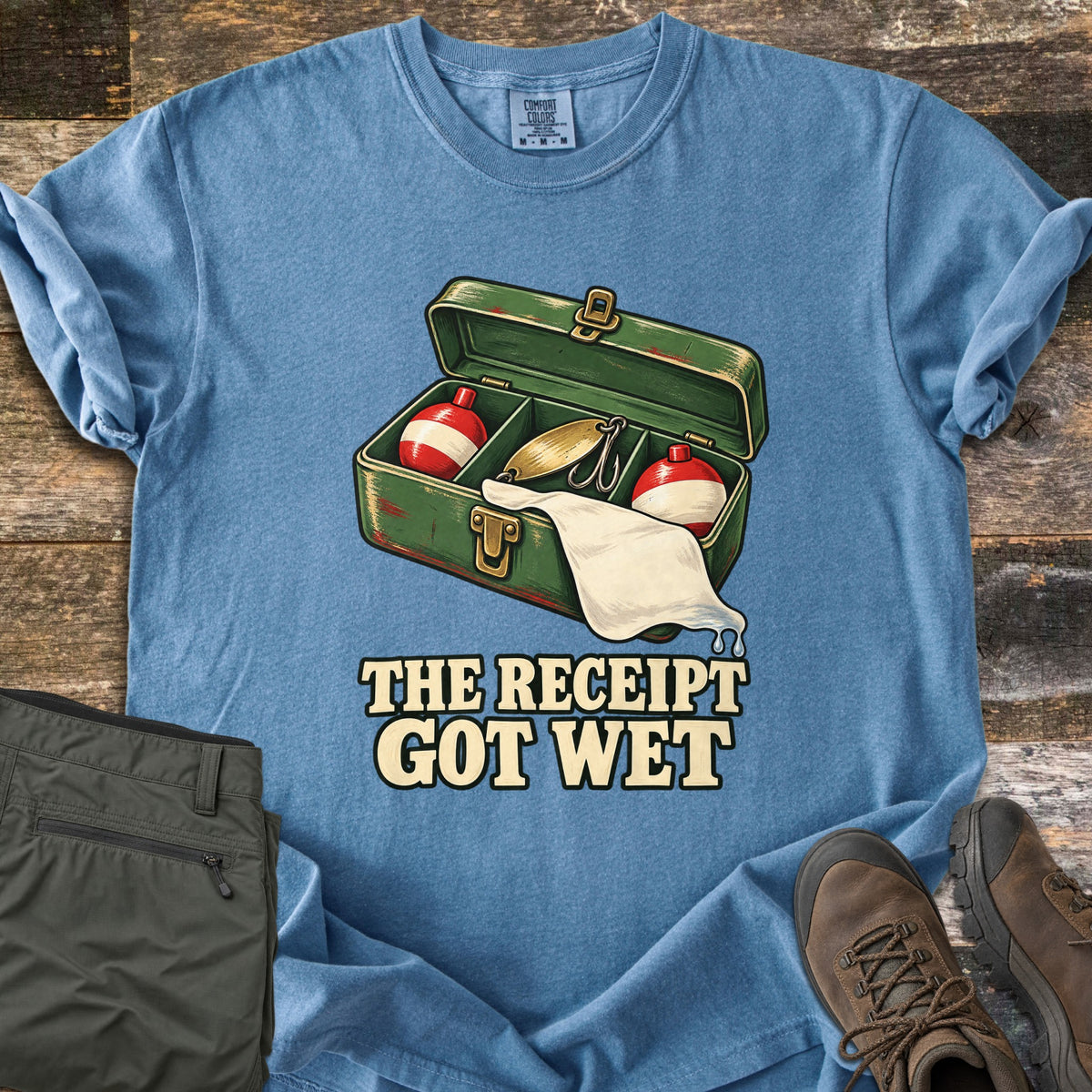 The Receipt Got Wet Comfort Colors T-shirt in Blue Jean, laid flat. An open green tackle box holds red and white bobbers and a gold spoon lure, with a dripping white paper hanging out, above the words The Receipt Got Wet. Olive pants and brown hiking boots sit on weathered wood.