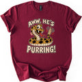Aww He's Purring Comfort Colors T-shirt in Maroon, laid flat. A grinning cartoon rattlesnake with a red forked tongue shakes its rattle between the words Aww, He's Purring! in cream letters. The shirt lies on a white background with cuffed sleeves.