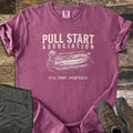 Pull Start Association Comfort Colors T-shirt in Berry, laid flat. Cream distressed text reads Pull Start Association above a sketched rowboat with an outboard motor on water, and below it It'll Start. Eventually. Sleeves are rolled on a wood floor beside green pants and brown hiking boots.