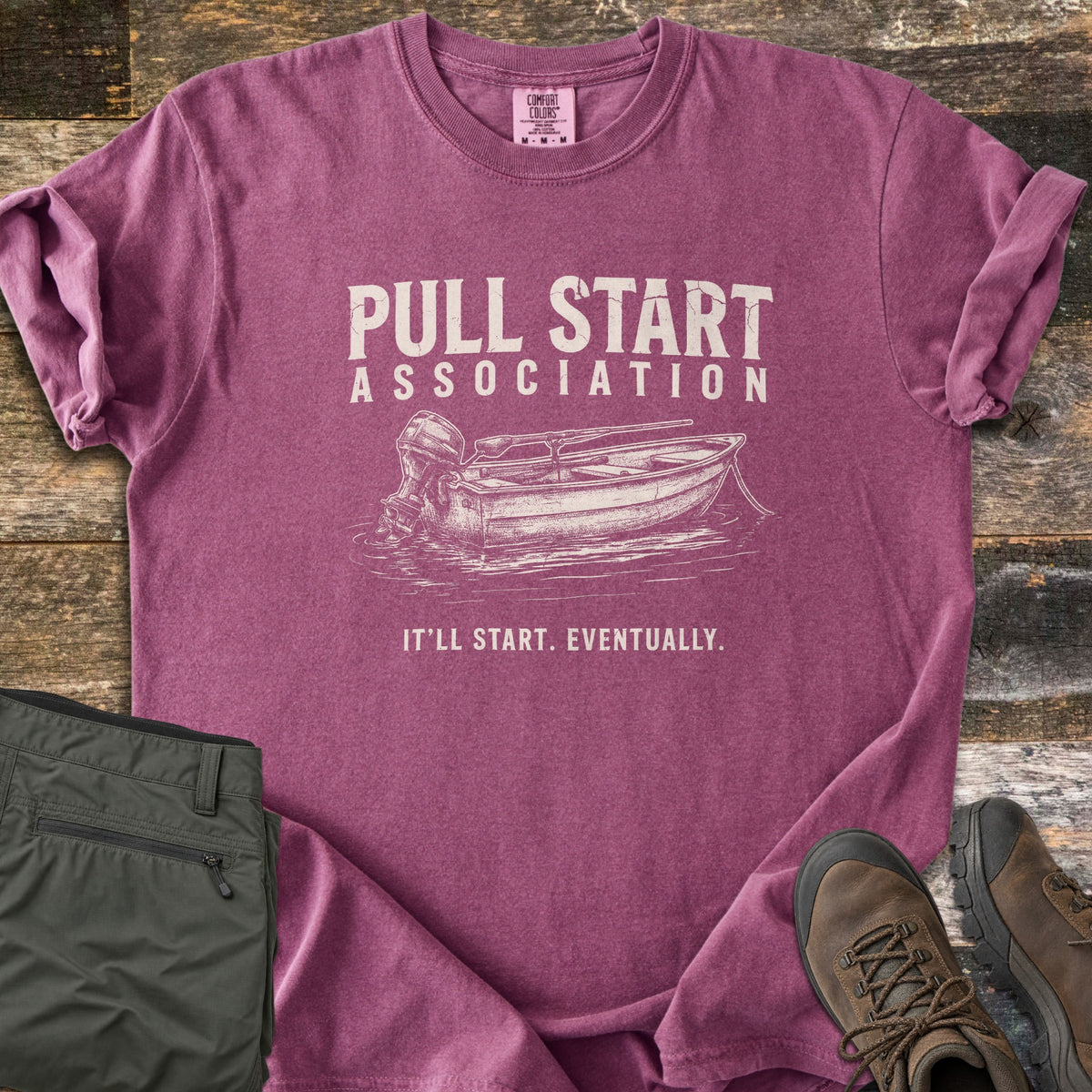 Pull Start Association Comfort Colors T-shirt in Berry, laid flat. Cream distressed text reads Pull Start Association above a sketched rowboat with an outboard motor on water, and below it It'll Start. Eventually. Sleeves are rolled on a wood floor beside green pants and brown hiking boots.