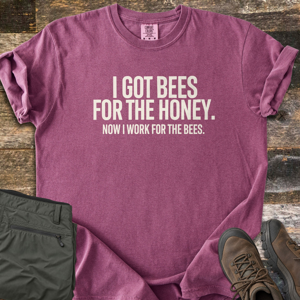 Work For The Bees Comfort Colors T-shirt in Berry, laid flat. Cream block letters read I GOT BEES FOR THE HONEY. NOW I WORK FOR THE BEES. The shirt lies with cuffed sleeves on weathered wood planks beside folded olive green pants and brown hiking boots.