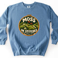 Moss in Stitches Comfort Colors crewneck sweatshirt in Blue Jean, laid flat. A wooden embroidery hoop frames stitched green moss mounds with two orange spore capsules, MOSS above and IN STITCHES below in cream yarn letters, and a needle with cream thread. On a white background.