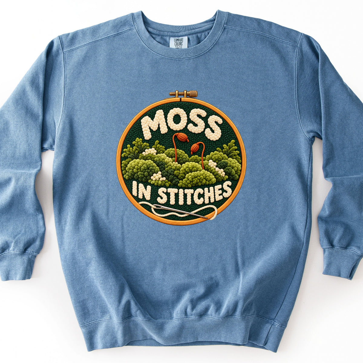 Moss in Stitches Comfort Colors crewneck sweatshirt in Blue Jean, laid flat. A wooden embroidery hoop frames stitched green moss mounds with two orange spore capsules, MOSS above and IN STITCHES below in cream yarn letters, and a needle with cream thread. On a white background.