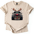 Santa Truck Comfort Colors T-shirt in Sand, laid flat. A front-facing dark gray pickup truck with reindeer antlers, a round red nose on its grille, and colored lights with a red bow on the bumper, amid snowflakes and red stars. No words. White background.