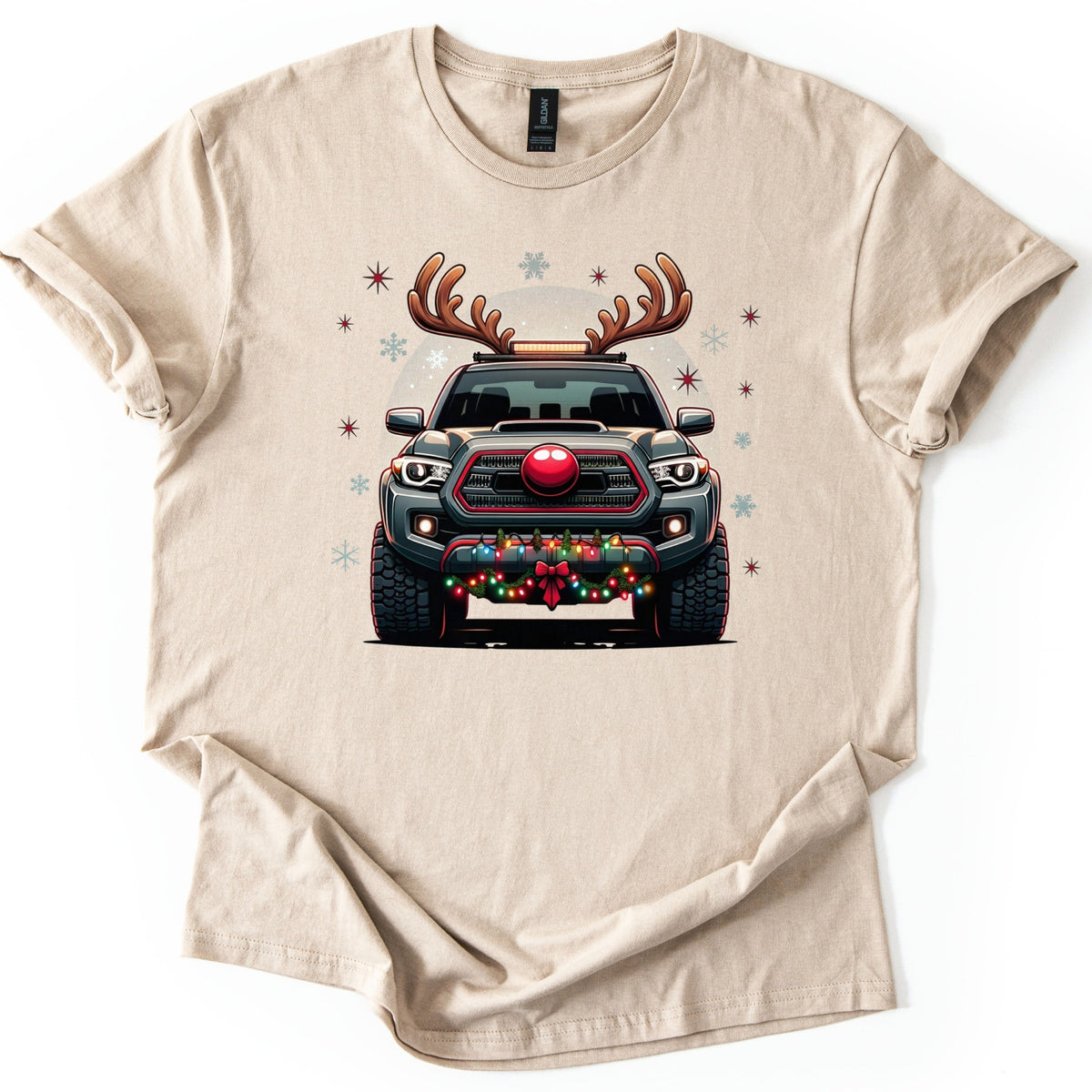 Santa Truck Comfort Colors T-shirt in Sand, laid flat. A front-facing dark gray pickup truck with reindeer antlers, a round red nose on its grille, and colored lights with a red bow on the bumper, amid snowflakes and red stars. No words. White background.
