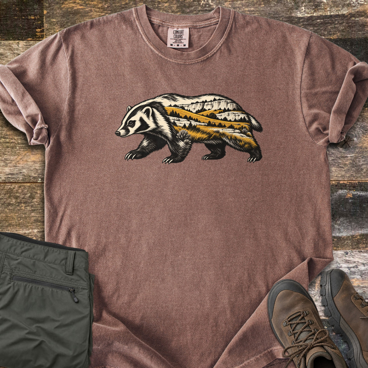 Badger Prairie Bluffs Comfort Colors T-shirt in Espresso, laid flat. A walking badger silhouette filled with a prairie scene of golden grass, pine trees and white bluffs is printed on the chest. Shown on weathered wood with folded olive pants and brown hiking boots.