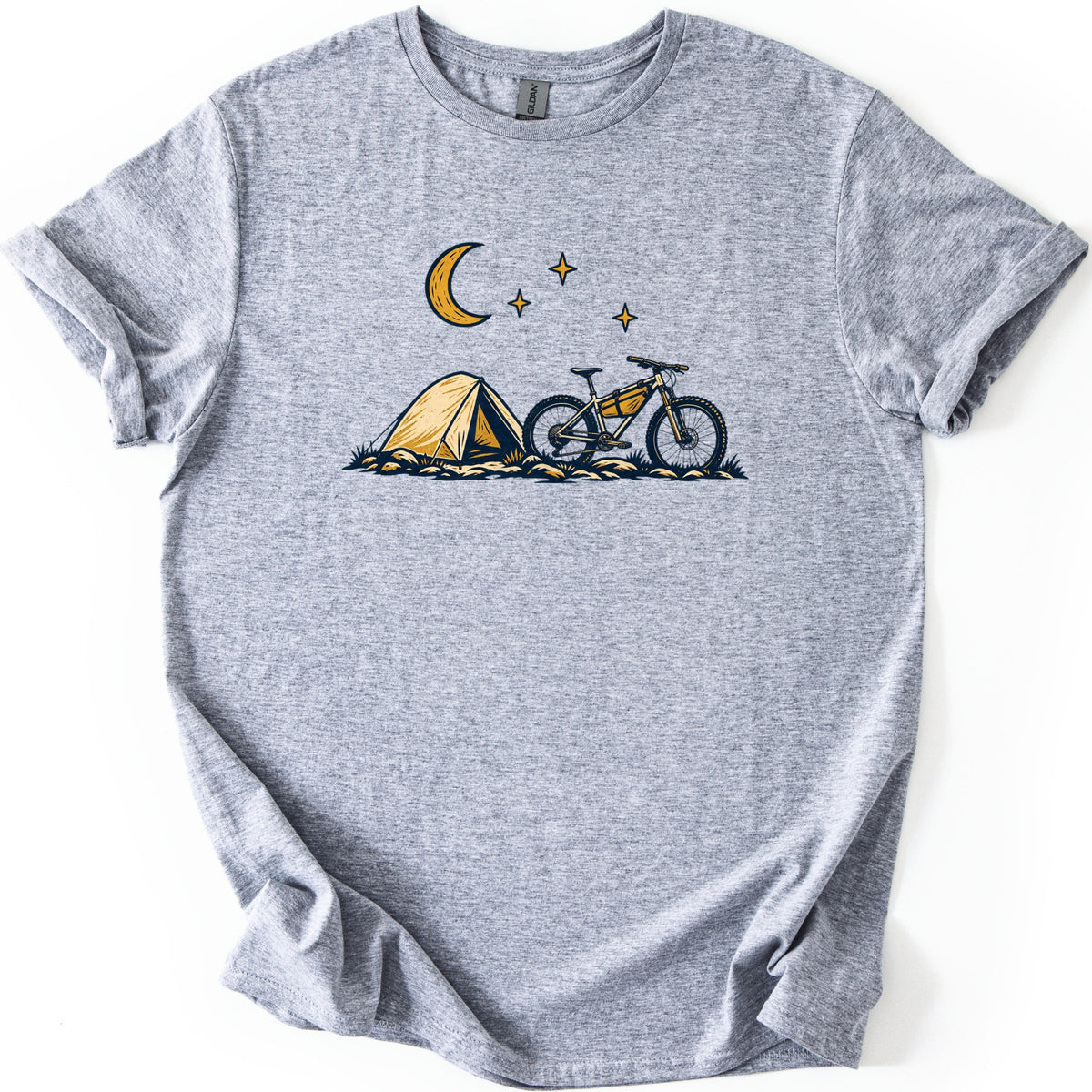 Bikepacking Under the Stars Comfort Colors T-shirt in Sport Grey, laid flat. A gold crescent moon and three stars shine above a yellow tent and a mountain bike with a frame bag on grassy, rocky ground. There are no words. The shirt has cuffed sleeves and lies on a white background.