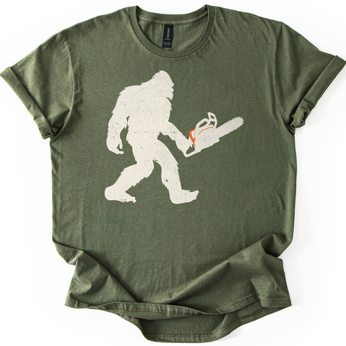 Lumber Squatch Comfort Colors T-shirt in Military Green, laid flat. A distressed cream silhouette of Bigfoot walking mid-stride while holding a chainsaw with an orange accent, with no words printed. The shirt has cuffed sleeves and lies on a white background.