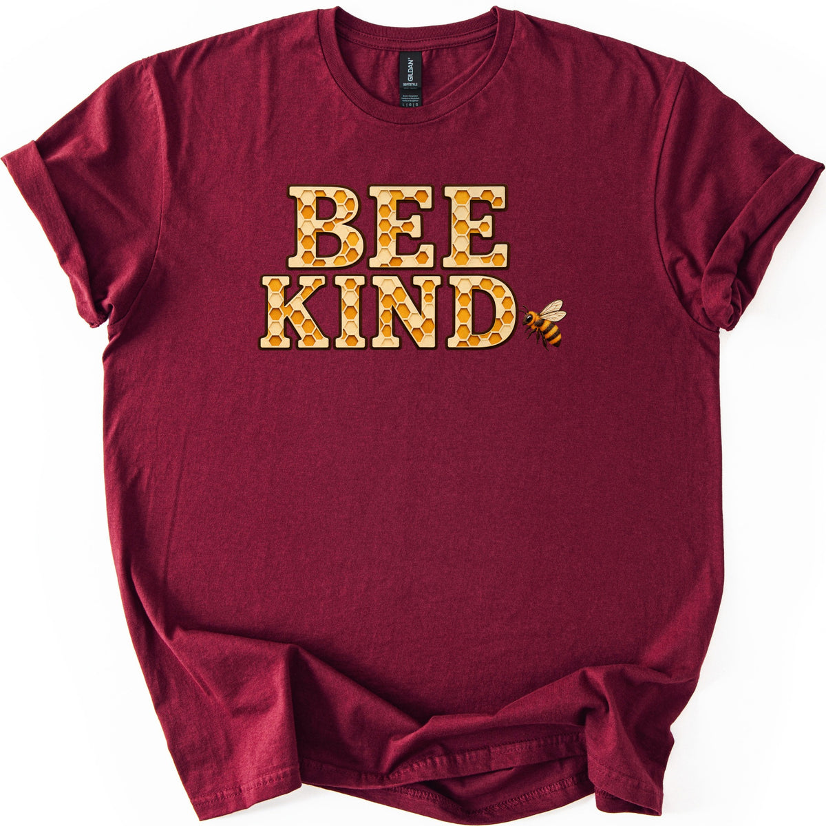 Bee Kind Comfort Colors T-shirt in Maroon, laid flat. The words BEE KIND in large block letters filled with a golden honeycomb pattern, with a small honeybee flying beside the D. The shirt has rolled sleeves and lies on a white background.
