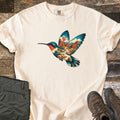 Hummingbird Canyon Comfort Colors T-shirt in Ivory, laid flat. A hummingbird in flight whose body and wings are filled with a desert canyon scene of red rock cliffs, a winding stream, boulders and red-blooming plants. Olive hiking pants and brown boots sit on weathered wood.