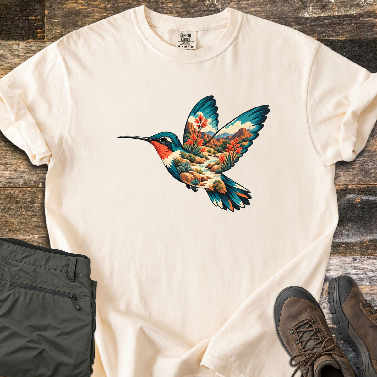 Hummingbird Canyon Comfort Colors T-shirt in Ivory, laid flat. A hummingbird in flight whose body and wings are filled with a desert canyon scene of red rock cliffs, a winding stream, boulders and red-blooming plants. Olive hiking pants and brown boots sit on weathered wood.