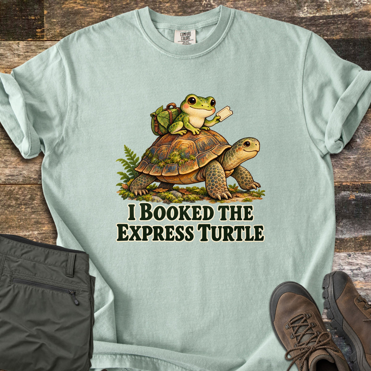 I Booked the Express Turtle Comfort Colors T-shirt in Bay, laid flat. A cartoon frog with a leafy backpack holds a ticket while riding a mossy tortoise, above the words I Booked the Express Turtle in dark green. Shown on weathered wood with folded olive pants and brown hiking boots.