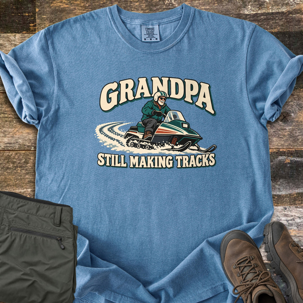 Grandpa Still Making Tracks Comfort Colors T-shirt in Blue Jean, laid flat. A cartoon grandpa in a helmet and sunglasses rides a vintage snowmobile through snow, with GRANDPA above and STILL MAKING TRACKS below. Olive pants and brown hiking boots sit beside it on weathered wood.
