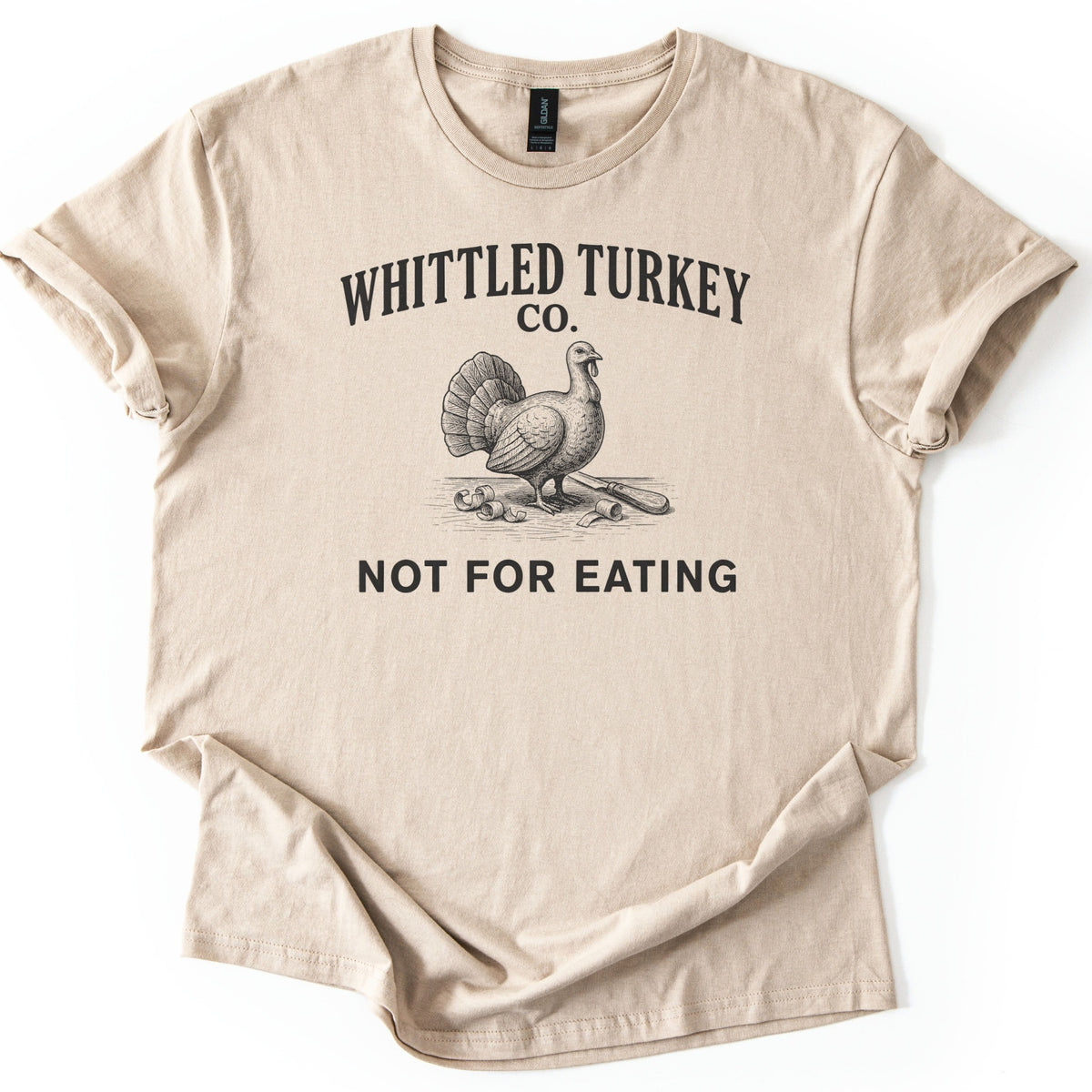 Whittled Turkey Co Comfort Colors T-shirt in Sand, laid flat. Black print of an engraved-style turkey beside wood shavings and a carving knife, under the words Whittled Turkey Co. and above Not For Eating. Sleeves are cuffed on a white background.