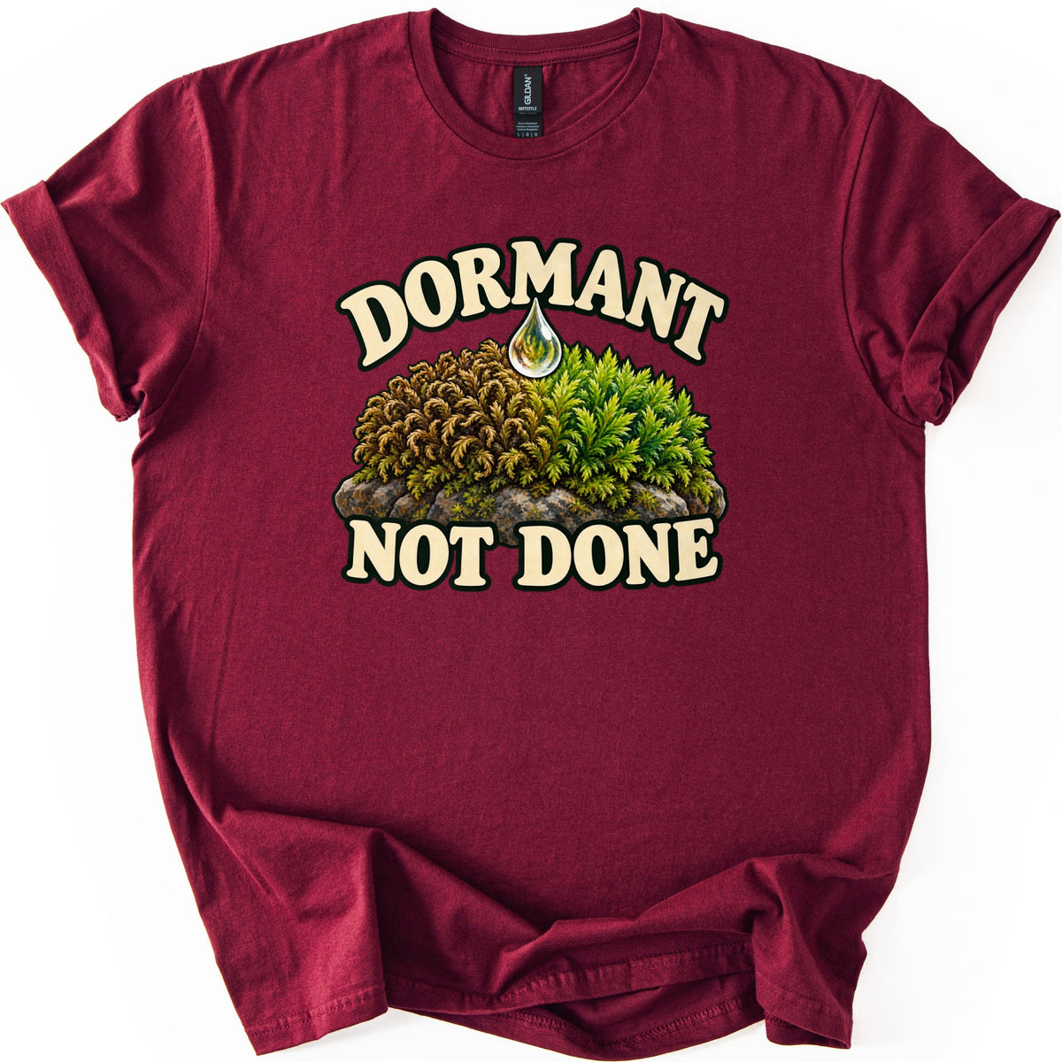 Dormant Not Done Comfort Colors T-shirt in Maroon, laid flat. A water drop falls onto a mossy rock, dry brown moss on the left and bright green on the right, between cream words Dormant above and Not Done below. Sleeves cuffed on a white background.