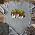 Built by Moss Comfort Colors T-shirt in Grey, laid flat. An illustrated chunk of brown soil topped with bright green moss and a small blue stream, with the words Built by Moss. Shown on weathered wood with folded olive hiking pants and brown hiking boots.