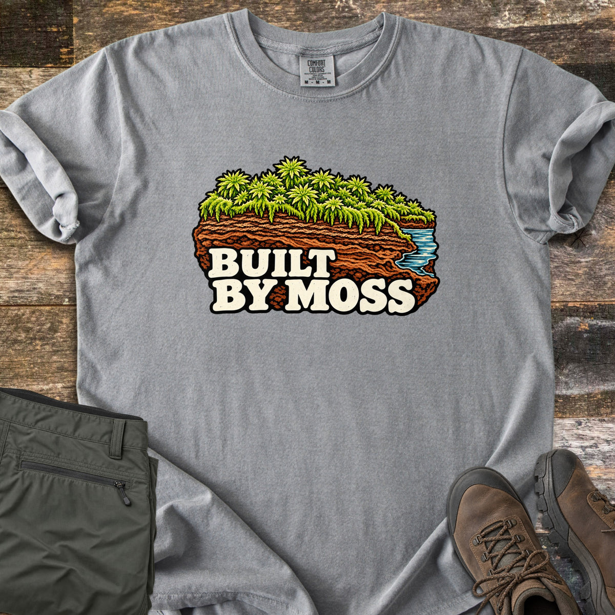 Built by Moss Comfort Colors T-shirt in Grey, laid flat. An illustrated chunk of brown soil topped with bright green moss and a small blue stream, with the words Built by Moss. Shown on weathered wood with folded olive hiking pants and brown hiking boots.