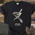 Small Bird Big Morning Comfort Colors T-shirt in Black, laid flat. A cream ink drawing of a small wren with a rust-colored breast perches on a seed-tipped twig above the words Small Bird. Big Morning. Olive hiking pants and brown hiking boots lie on weathered wood beside it.