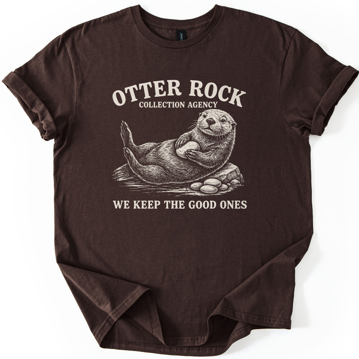Otter Rock Collection Agency Comfort Colors T-shirt in Dark Chocolate, laid flat. A cream sketch of a sea otter lounging on a rock, hugging a smooth stone beside a pile of pebbles, under the words Otter Rock Collection Agency and above We Keep the Good Ones. Sleeves cuffed, on a white surface.