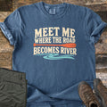 Meet Me Where the Road Becomes River Comfort Colors T-shirt in Denim, laid flat. Cream text reads Meet Me Where the Road Becomes River, with an orange canoe paddle between the lines and turquoise water swirls below. Sleeves are rolled on wood planks beside olive hiking pants and brown boots.